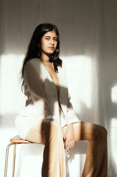 The singer Naari is sitting on a stool. She is wearing a beige blazer and beige pants looking directly into the camera. 