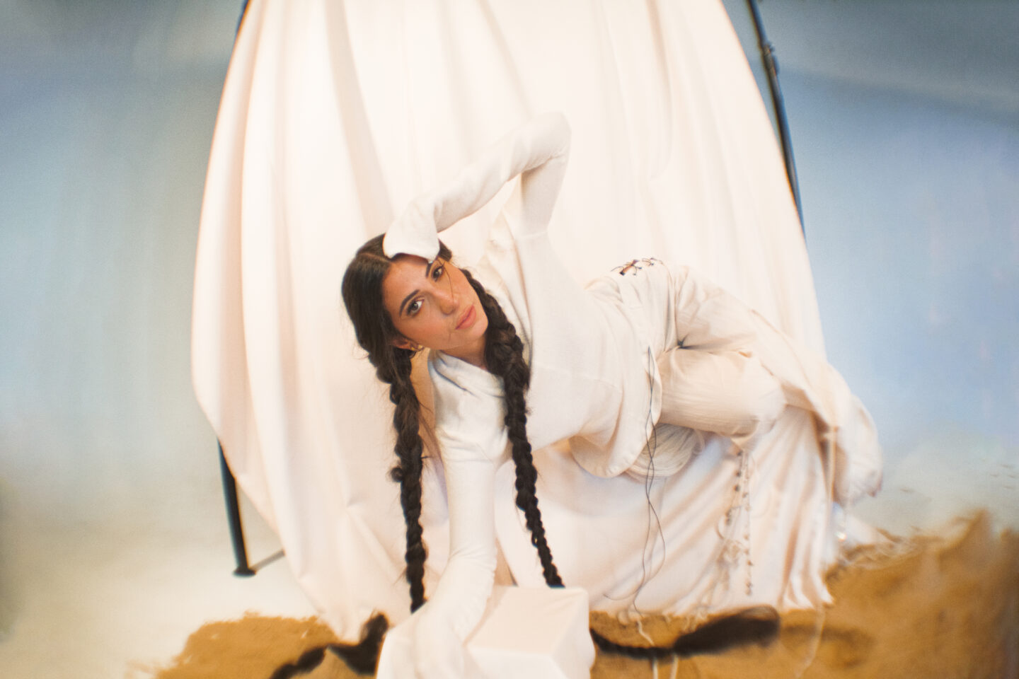 Singer Evin is reclining on a chair. Dressed in white against a white cloth backdrop. Her hair is braided she looks into the camera.