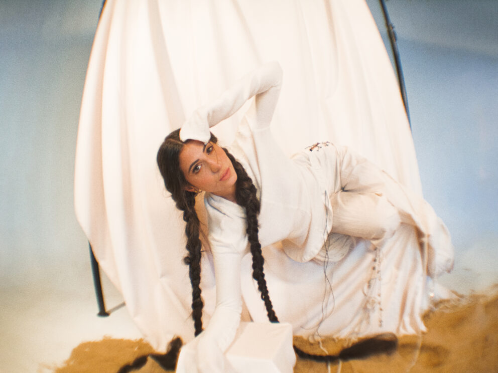 Singer Evin is reclining on a chair. Dressed in white against a white cloth backdrop. Her hair is braided she looks into the camera.