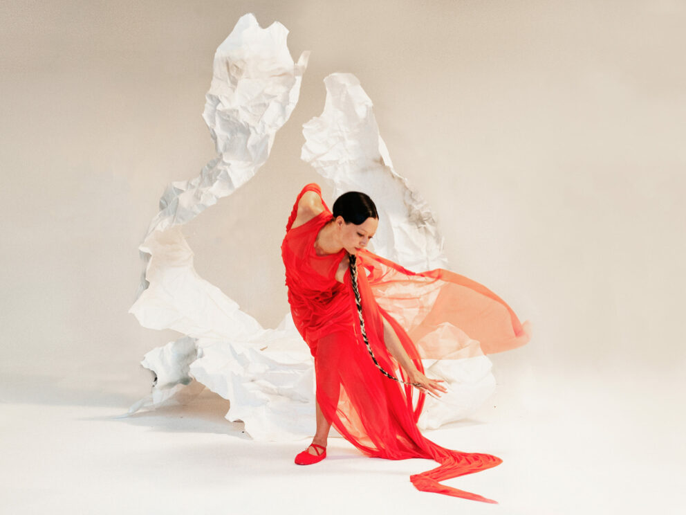 A wide shot of Lucina Chua dressed in a red dress dancing in front of a white abstract sculpture in a white studio setting.