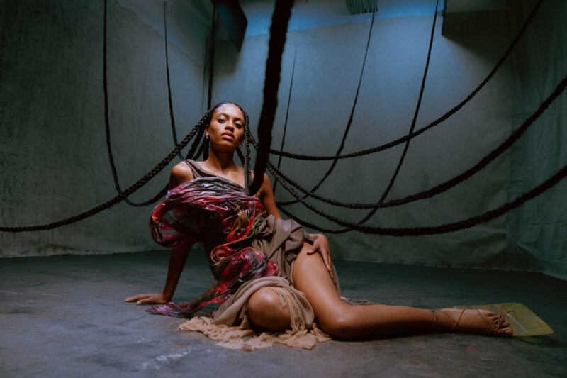 In a wide shot Annahstasia sits in the middle of the room. Her long braids float upward and out of the frame. she is wearing a textured dress made of red and purple fabric.