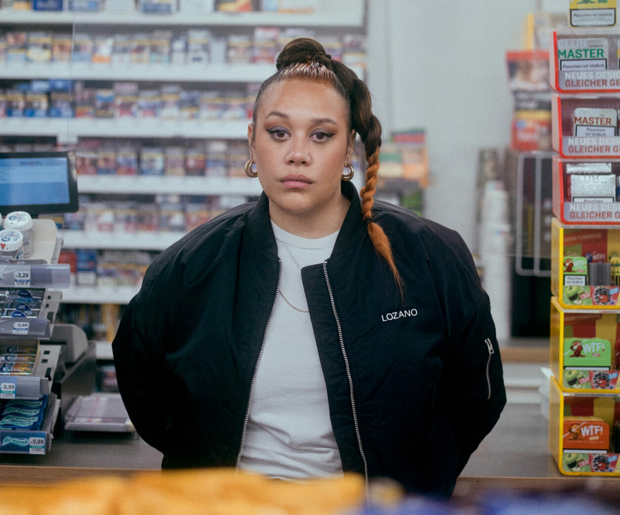 Singer Ray Lozano is standing in a convenience store. Taken from behind the counter, we see the singer from the waist up. She is looking directly at the camera wearing a black bomber jacket, a white t-shirt, and her hair tied into a high ponytail.