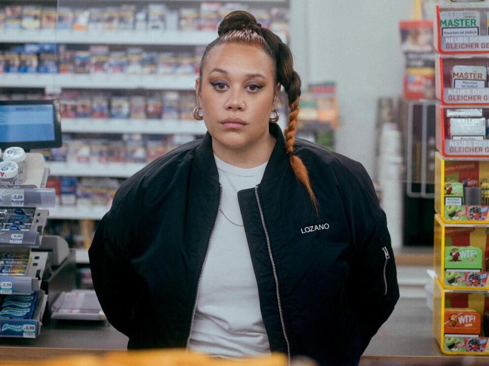 Singer Ray Lozano is standing in a convenience store. Taken from behind the counter, we see the singer from the waist up. She is looking directly at the camera wearing a black bomber jacket, a white t-shirt, and her hair tied into a high ponytail.