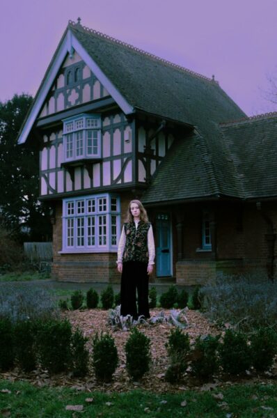 Neev stands in front of a retro style country house. The colors are lilac tinted and she is looking directly into the camera.  