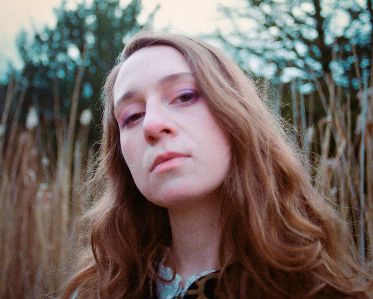 Portrait shot of Neev. She looks into the camera. Her blond her flows over her shoulders. She is standing in nature. in the background are trees.