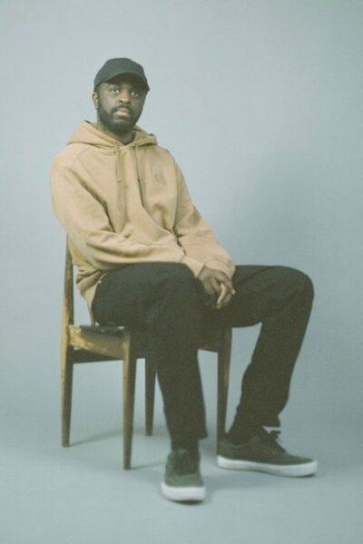 Alfa Mist sits in a chair in front of a white background. He is looking directly into the camera wearing a yellow hoodie and black pants. The image has a greenish hue. 
