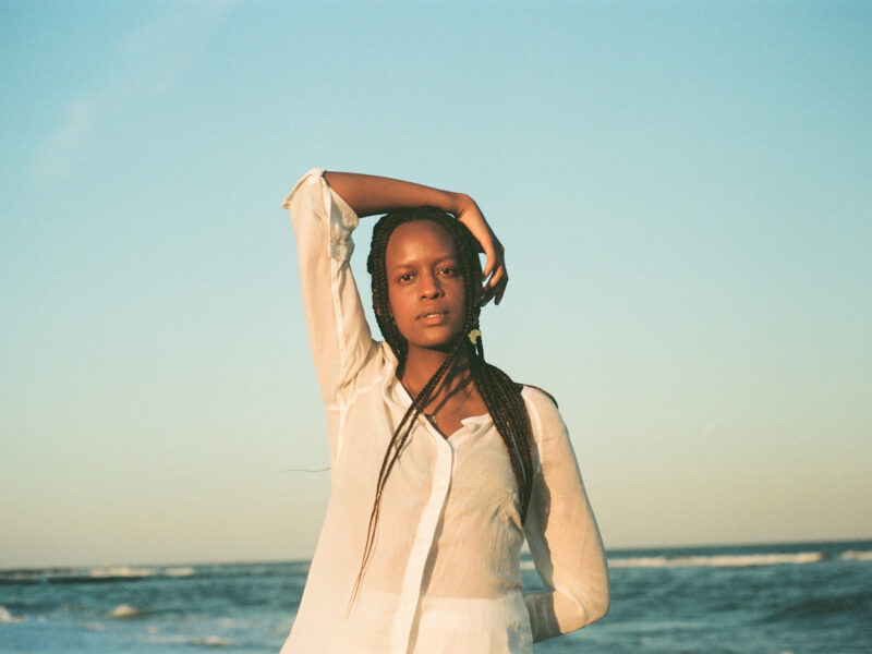 Nyokabi Kariuki is standing in front of the ocean. She is wearing a white top and has one arm resting on her head. Her hair is braided and falls over her shoulder.