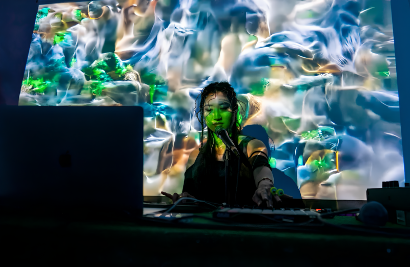 The artist at a live show. She is sitting behind a desk and laptop. Projections of green-blue water textures cover the stage.