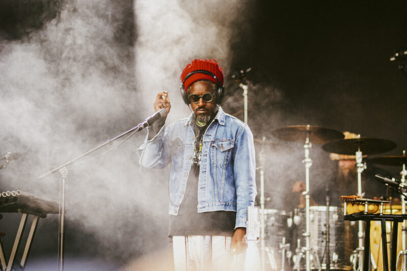 André 3000 on stage holding the microphone. behind him the stage is hazy. he is looking down, wearing sunglasses and a red hat