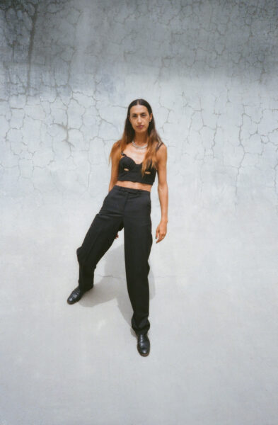 A woman in black suit pants and a black crop top is standing in front of a concrete wall.