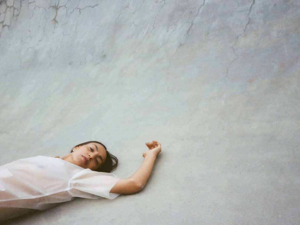 A woman lying on concrete floor with her arm stretched above her heard. She is in the bottom left corner of the image. Her legs are cropped out.