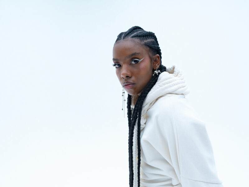 A dark-skinned person looking into the camera in front of a white backdrop wearing a white hoodie and long braids.