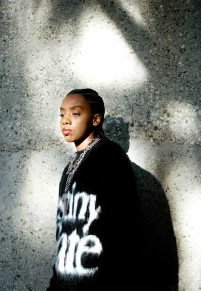 A dark-skinned person standing in front of a wall looking away wearing a large black sweatshirt with white letters on it and several silver chains